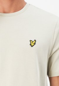 Beige bomulds-T-shirt med rund halsudskæring, prydet med en lille sort og gul fuglebroderi på venstre bryst. Glat tekstur.