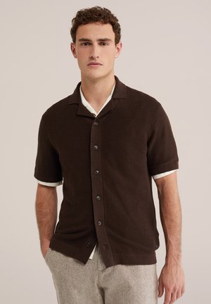 Mężczyzna ubrany w ciemnobrązową koszulę z krótkim rękawem na guziki, na białej koszulce, w jasnoszarych spodniach, stojący na jednolitym tle.