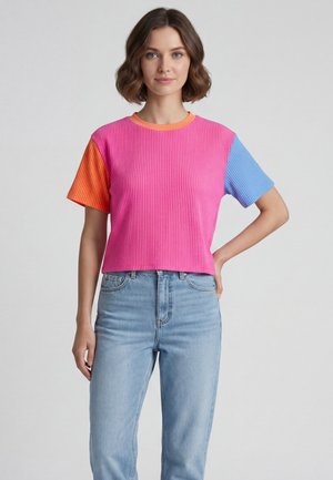 Kvinde med kort bølget hår står med den ene hånd på hoften iført en pink ribbet t-shirt med orange og blå ærmer og lyseblå jeans.