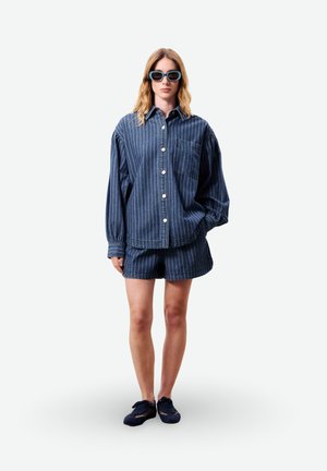 Jeune femme portant une chemise boutonnée à rayures bleues surdimensionnée, un short assorti, des baskets foncées et des lunettes de soleil bleues, debout devant un fond blanc.