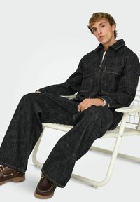 Melns džinsa kombinezons ar garām piedurknēm, priekšā rāvējslēdzēju, divām krūšu kabatām un platām biksēm, kombinēts ar brūnām šņorzābakiem.