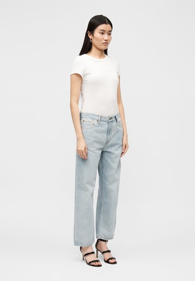 Γυναίκα που στέκεται, φοράει εφαρμοστό λευκό κοντομάνικο τοπ, ανοιχτό μπλε ψηλόμεσο τζιν, χρυσούς κρίκους σκουλαρίκια και μαύρα σανδάλια με τακούνι.