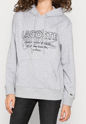 Personne portant un sweat à capuche Lacoste gris clair avec un crocodile dessiné en noir et du texte, associé à un pantalon noir.