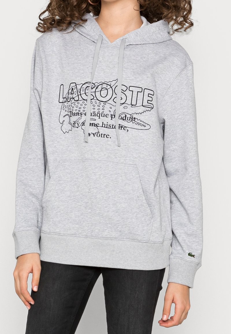 Personne portant un sweat à capuche Lacoste gris clair avec un crocodile dessiné en noir et du texte, associé à un pantalon noir.