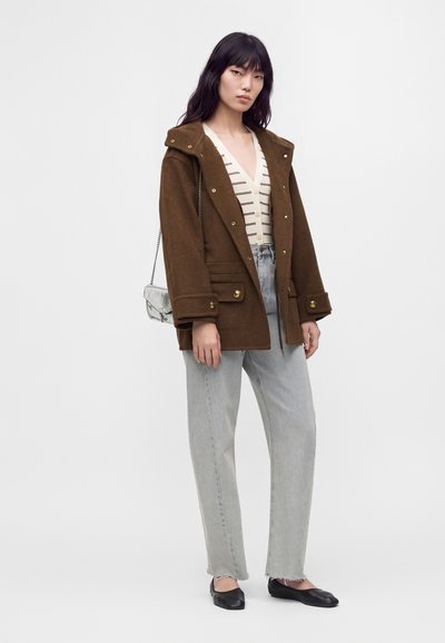 Jeune femme portant un manteau marron, un cardigan blanc rayé, un jean gris clair, des ballerines noires et portant un petit sac à bandoulière gris, debout de manière neutre.