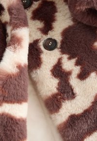 Tkanina z futra sztucznego o kremowo-brązowym wzorze przypominającym krowę, z okrągłymi czarnymi guzikami i miękkim, teksturowanym wykończeniem.