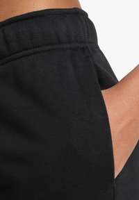 Hånd inne i frontlommen på svarte elastiske bukser med strikk i livet, som viser tekstur av stoffet og detaljene i sømmen.