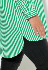 Camisa a rayas verdes y blancas con dobladillo curvado y puño abotonado, combinada con leggings oscuros. Presenta un material de algodón nítido.