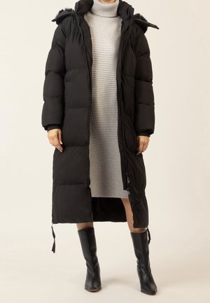 Vrouw draagt een lange zwarte donsjas over een lichtgrijze geribbelde coltrui jurk en zwarte laarzen tot de knie, staand tegen een effen achtergrond.