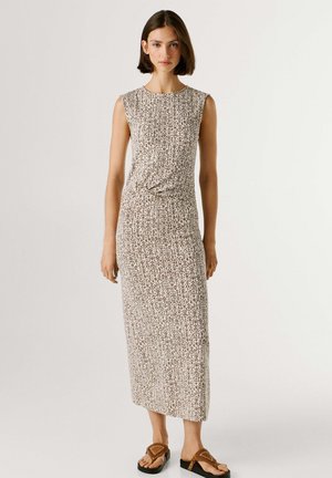 Mujer con cabello corto castaño que lleva un vestido beige sin mangas hasta el tobillo con un sutil patrón abstracto y sandalias marrones.