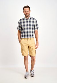 Camicia a maniche corte a quadri in blu e crema, abbinata a pantaloni cargo beige con tasche laterali e scarpe da ginnastica grigie.