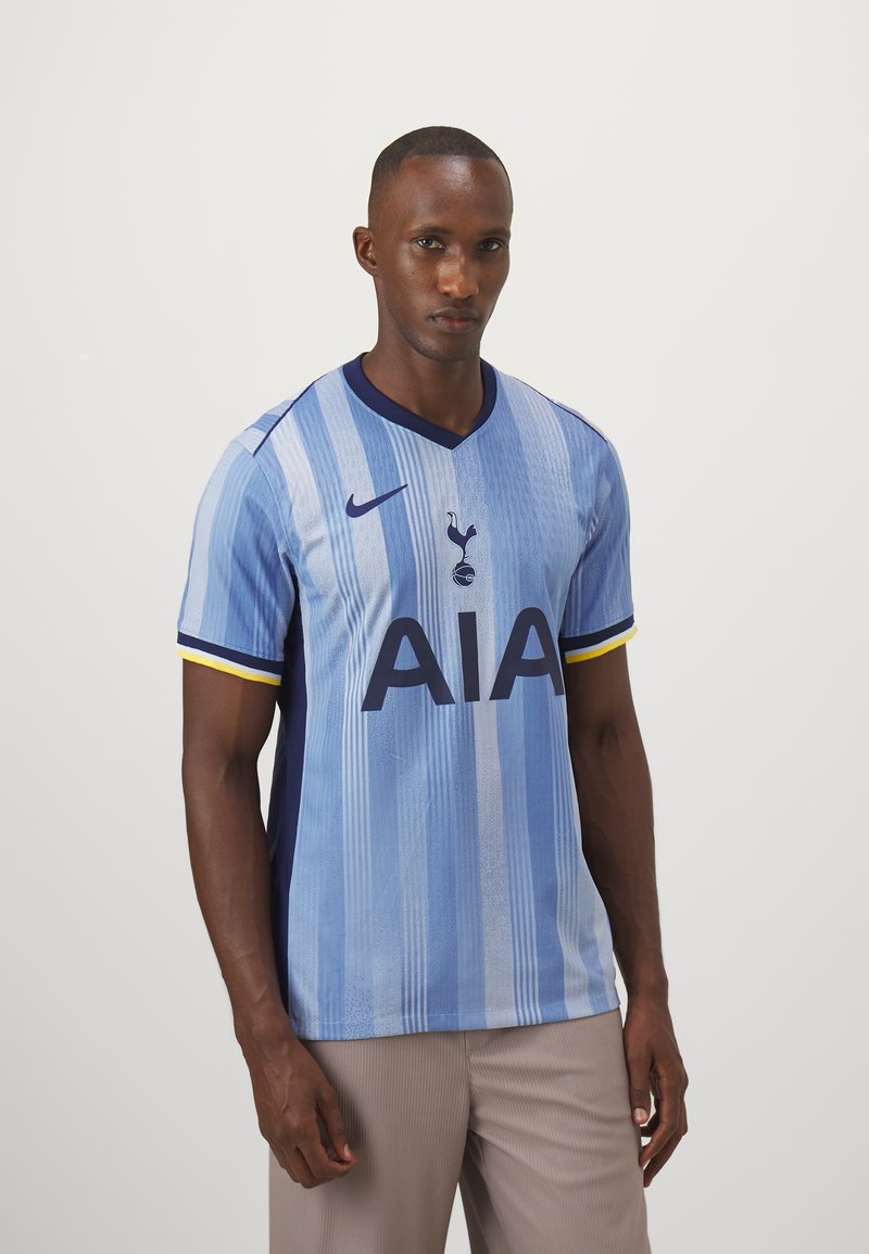 Mężczyzna w jasnoniebieskiej, w paski koszulce piłkarskiej Tottenham Hotspur z granatowymi i żółtymi akcentami, w beżowych spodniach, stojący na jednolitym tle.