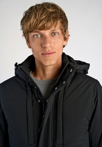 Mustat eristetyt takki hupulla, jossa on vetoketju ja kiristysnauha. Päällä harmaa teksturoitu neule, tiukasti istuva malli.