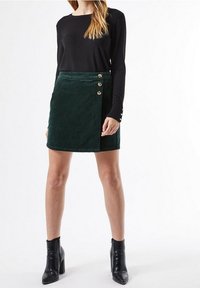 Grön kordfläsk minikjol med omlottdesign och tre guldknappar på framsidan. Kombineras med en svart långärmad topp och svarta ankelboots.