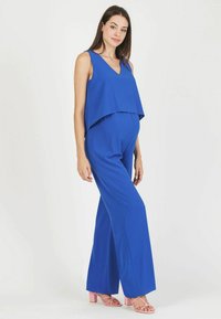 Mono azul con escote en V y parte superior fluida, con piernas anchas. Llevado con sandalias de tacón cuadrado rosas, de textura suave y diseño casual.