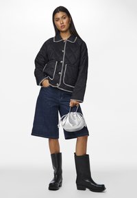 Giacca nera trapuntata con dettagli in cucitura bianca, due tasche frontali, abbinata a pantaloni bermuda in denim scuro e stivali neri alti fino al ginocchio, mentre tiene una borsa argentata.