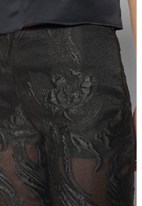 Primer plano de pantalones negros transparentes con bordado floral intrincado, mostrando la textura de la tela y parte de una blusa oscura.