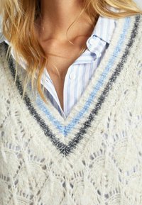 Pull en maille crème avec un motif en dentelle détaillé, doté d'un col en V profond et d'une bordure multicolore en gris et bleu clair. Chemise rayée en dessous.