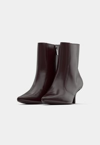 Pár tmavohnedých kožených členkových čižiem s špicatými špičkami, bočnými zipsami a kitten podpätkami na jednoduchom bielom pozadí.