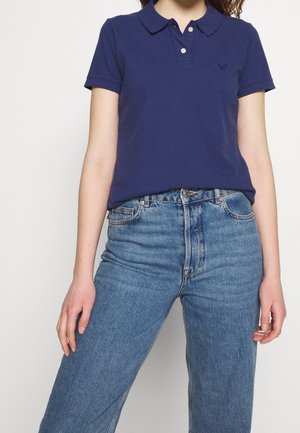 Kobieta ubrana w granatową koszulkę polo z krótkim rękawem i niebieskie wysokie dżinsy, stojąca na białym tle.