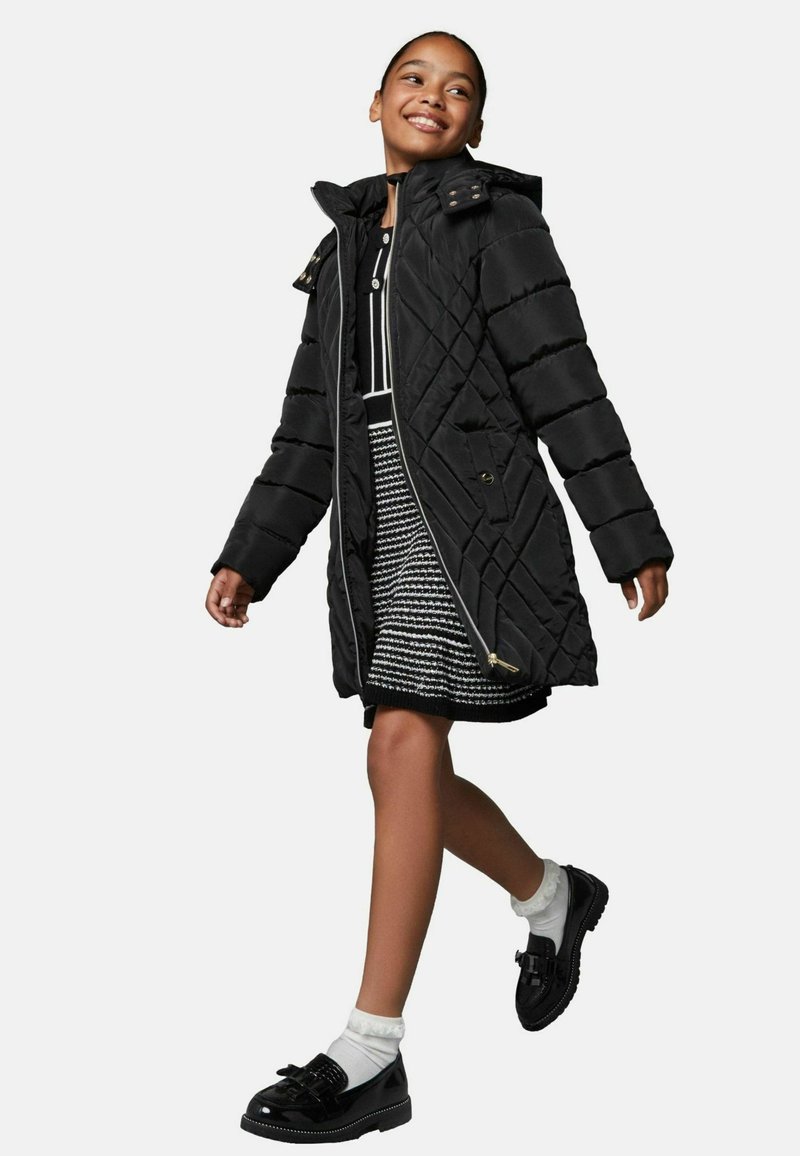 Zwarte gewatteerde pufferjas met een capuchon, ritssluiting en gouden accenten. Draagt over een zwart-wit patroonjurk en gecombineerd met zwarte schoenen.