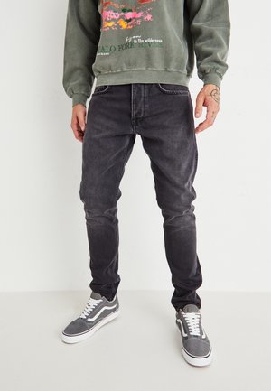 Persona con jeans neri sbiaditi, sneaker grigie e bianche, e una felpa verde con testo e grafiche colorate, in piedi davanti a uno sfondo bianco.