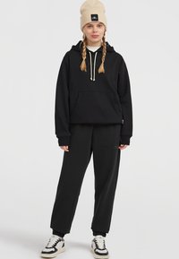 Sort hættejakke med frontlomme, snører og logo; kombineret med sorte sweatpants og multicolore sneakers. Beige strikket hue.