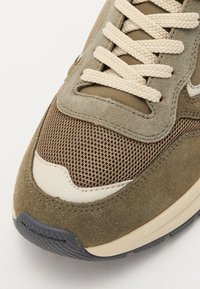 Sneaker in suede marrone con dettagli in rete, rifiniture color crema e suola in gomma testurizzata. Caratterizzato da lacci beige e una forma slanciata.