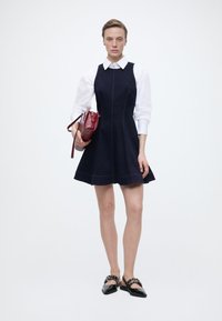 Γυναίκα που φοράει εφαρμοστό μπλε-σκούρο φόρεμα πάνω από λευκό πουκάμισο, κρατώντας κόκκινη τσάντα χειρός και φορώντας μαύρες μυτερές μπαλαρίνες με λουράκια και τρουκς.