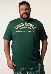 Zelené bavlněné tričko s potiskem "GOLD COAST, QUEENSLAND" a "CHASING WAVES SINCE 2007" v béžové barvě. Krátké rukávy a kulatý výstřih.