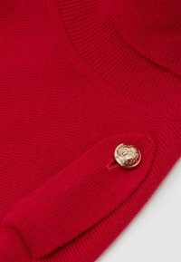 Maglione rosso a maglia con colletto a costine, dettaglio di bottoni laterali e un bottone decorativo dorato con un design in rilievo.