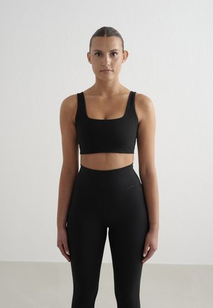 Young woman with hair tied back wearing black sports bra and high-waisted leggings, standing against a plain light background.