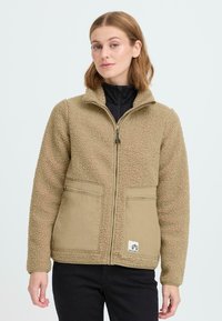 Tan fleece jakke med en tekstureret overflade, lynlås foran, to frontlommer og et let tan stofoverlay på de nedre sektioner.