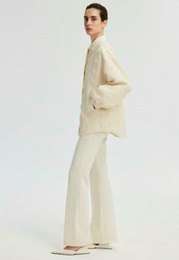 Veste texturée de couleur crème, à coupe ample et fermeture à boutons. Associée à un pantalon blanc taille haute, évasé, et des ballerines pointues.