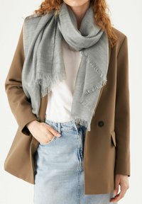 Lichtgrijze geweven sjaal met franjes, om de nek gedrapeerd, gecombineerd met een bruine blazer en witte shirt, voor een lichte achtergrond.