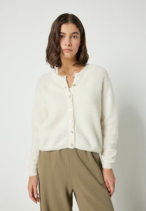 Giovane donna con capelli scuri alla lunghezza delle spalle che indossa un cardigan bianco con bottoni e pantaloni marroni larghi, su uno sfondo semplice.