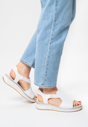 White open-toe sandals with cut-out sides, a cushioned footbed, grey sole, and light brown rubber outsole. Worn with light blue jeans.