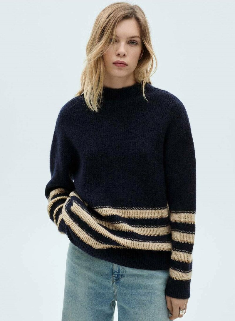 Maglione a collo alto di colore blu navy con orlo a righe beige e blu navy. Texture morbida e vestibilità rilassata. Indossato con jeans azzurri.