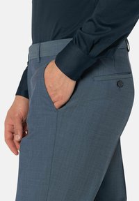 Man in een donkerblauw shirt en lichtblauwe broek, met één hand in de voorzak, waar de zakdetails en de knoopzak aan de achterkant zichtbaar zijn.