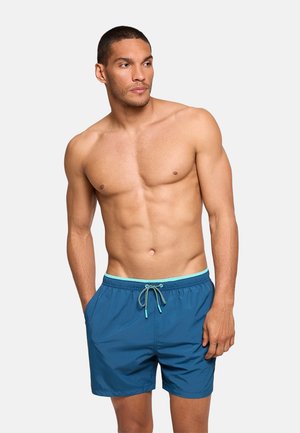 Hombre sin camiseta con cabello corto que lleva bañador azul con cordón, de pie con las manos en los bolsillos mirando hacia un lado.