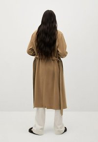 Langer beigefarbener Trenchcoat aus weichem Stoff mit einem Wickeldesign und einem passenden Gürtel. Getragen über weißen Hosen und schwarzen Sandalen.