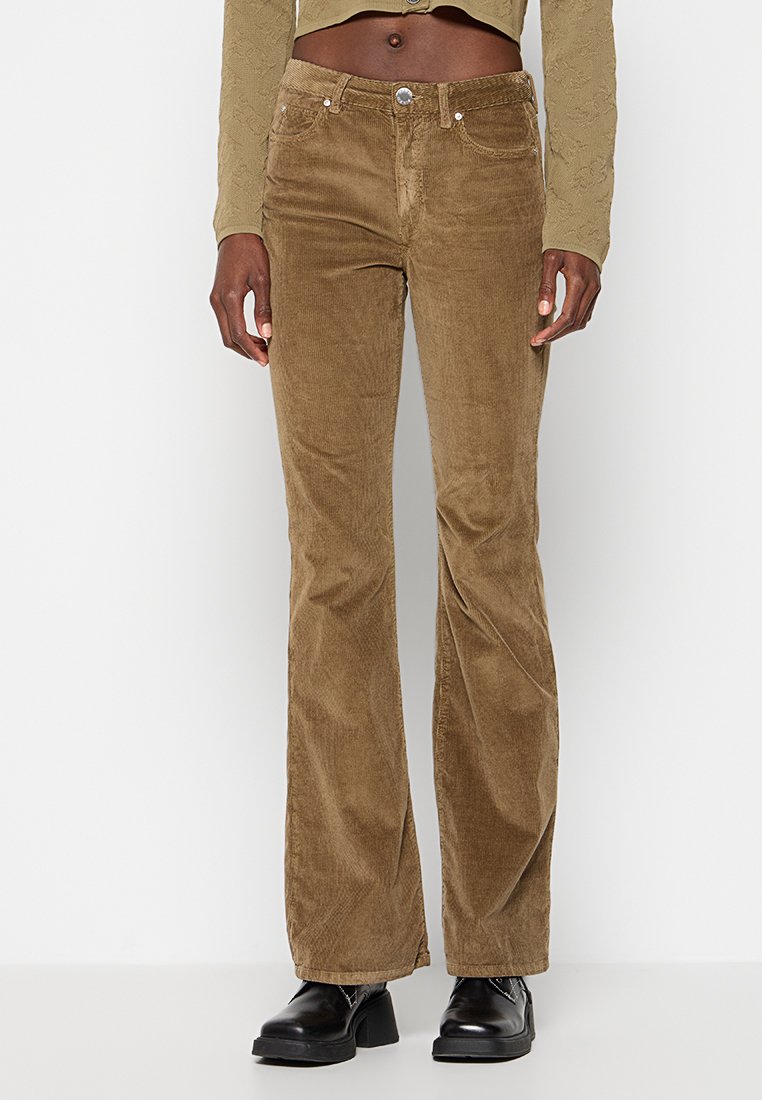 Osoba ubrana w brązowe spodnie sztruksowe w fasonie dzwonów, krótki beżowy top z długim rękawem i czarne buty na platformie na jednolitym tle.