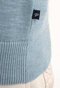 Světle modrý pletený svetr s ribbeným lemem a malou tmavou etiketou s bílým nápisem na boku. Měkká textura, klasický střih.