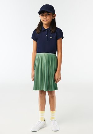 Granatowa koszulka polo, plisowana zielona spódnica, białe trampki i skarpetki w żółte paski. Czapka dopasowana kolorystycznie do koszulki. Widoczne teksturowane plisowania.