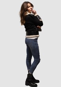 Mujer con chaqueta de piel de oveja negra, vaqueros ajustados azules con adornos laterales y botas de plataforma negras posando de lado contra un fondo liso.