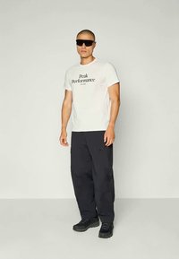 Witte t-shirt met de tekst "Peak Performance", zwarte cargo broek en zwarte sneakers. Model draagt donkere zonnebril. Eenvoudige achtergrond.