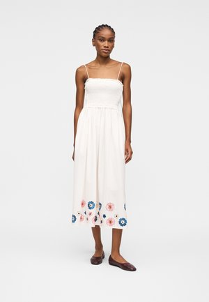 Donna in piedi che indossa un abito midi bianco senza maniche con corpetto arricciato e motivo floreale vicino all'orlo, abbinato a scarpe piatte scure.