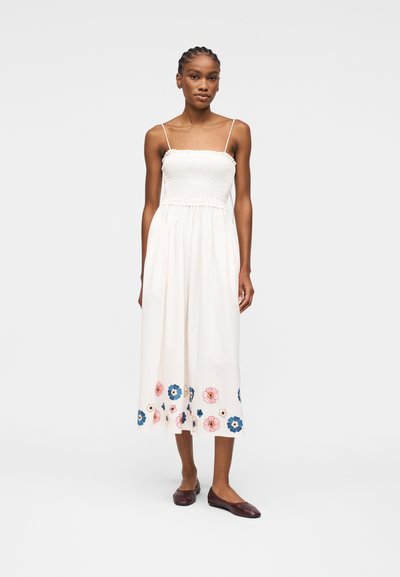 Femme debout portant une robe midi blanche sans manches avec un corsage smocké et un motif floral près de l'ourlet, associée à des chaussures plates foncées.