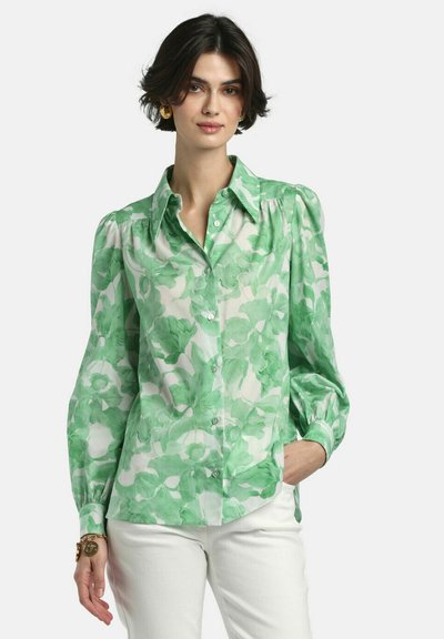 Sieviete zaļā ziedu raksta pogām aizpogātā blūzē ar uzpūstām piedurknēm, baltās biksēs, zelta auskariem un rokassprādzēm, stāvot ar vienu roku kabatā.