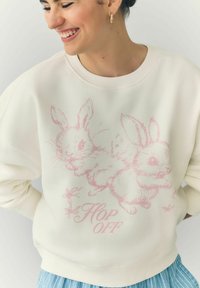 Creamfarbener Kapuzenpullover mit einer pinken Illustration von zwei Kaninchen. Der Text "HOP OFF" ist enthalten, mit geripptem Kragen und Bündchen.